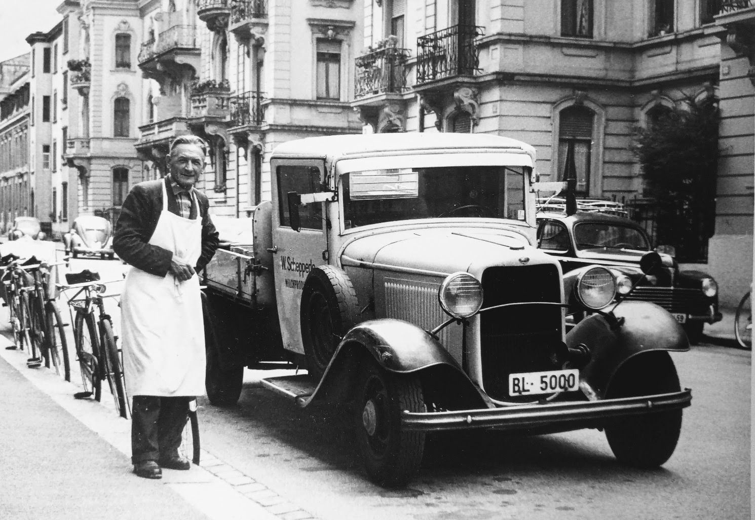 Milchmann mit weisser Schürze und Schnurrbart neben einem Flachbettlastwagen aus den 1930er Jahren mit dem Kennzeichen BL 5000, vor Basler Belle-Époque-Fassaden