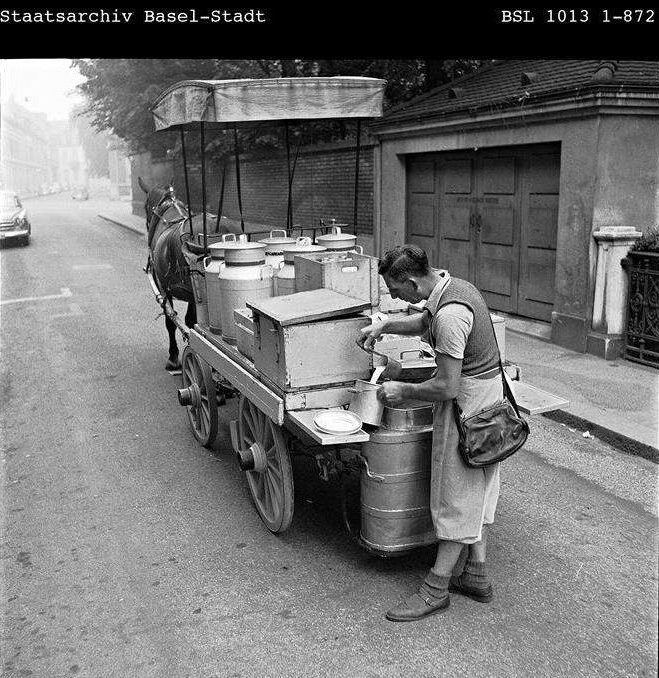 Milchmann mit Schürze und Schultertasche schöpft Milch aus einer Kanne in das Gefäss eines Kunden, am Pferdekarren auf einer Basler Strasse